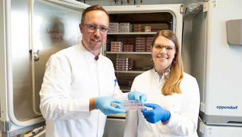 Ein Mann und eine Frau stehen in einem Labor vor einem geöffneten Schrank. Sie halten eine Laborschale in die Kamera.