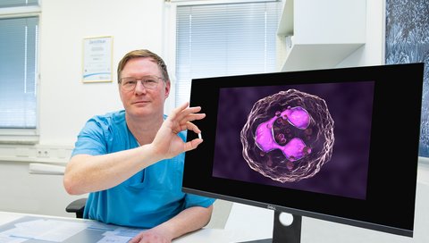 Ein Mann im blauen Kittel sitzt neben einem Bildschirm mit der Abbildung einer Immunzelle und hält eine weiße Tablette in der Hand.