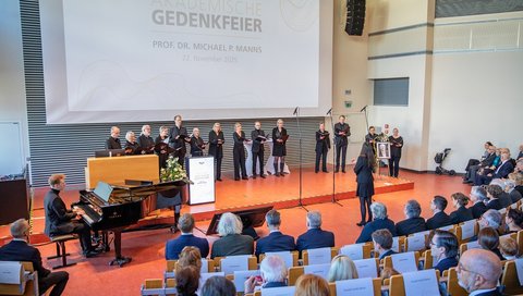 Im Hörsaal F der MHH steht auf dem Podium der MHH-Chor, daneben ein Mann am Klavier, in den Sitzreihen sitzen viele Menschen. 