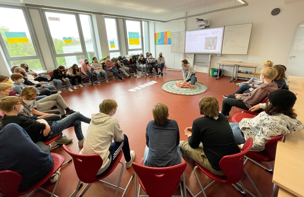 Schüler:innen sitzen im Kreis und in der Mitte erklärt ein Studierender die Grundlagen der Wiederbelebung. 