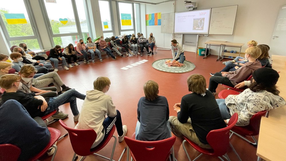 Schüler:innen sitzen im Kreis und in der Mitte erklärt ein Studierender die Grundlagen der Wiederbelebung. 