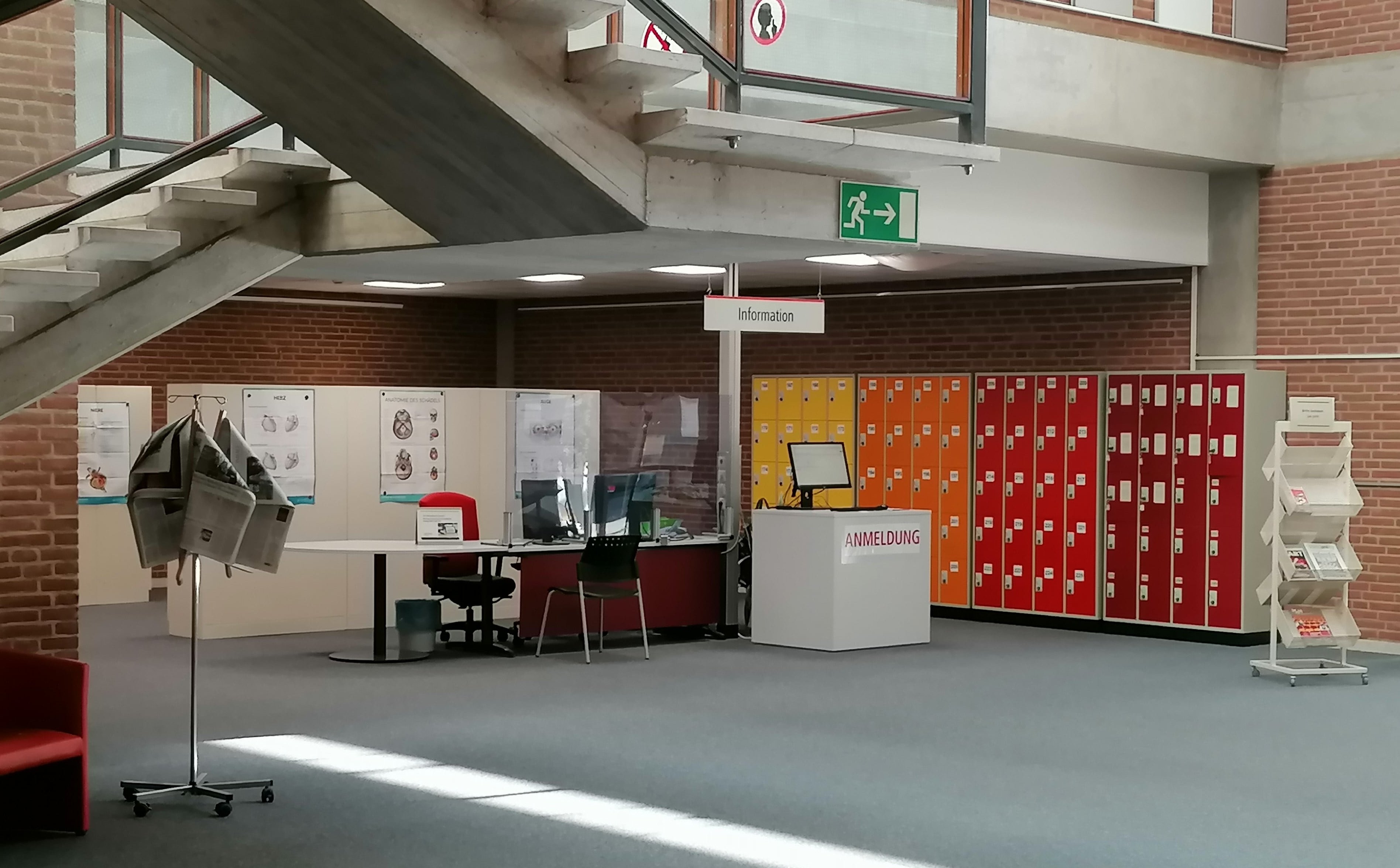 Foyer der Bibliothek mit Schreibtisch der Information und den Schließfächern im Hintergrund, links roter Sessel und Ständer mit Tageszeitungen, rechts Ständer mit Info-Material