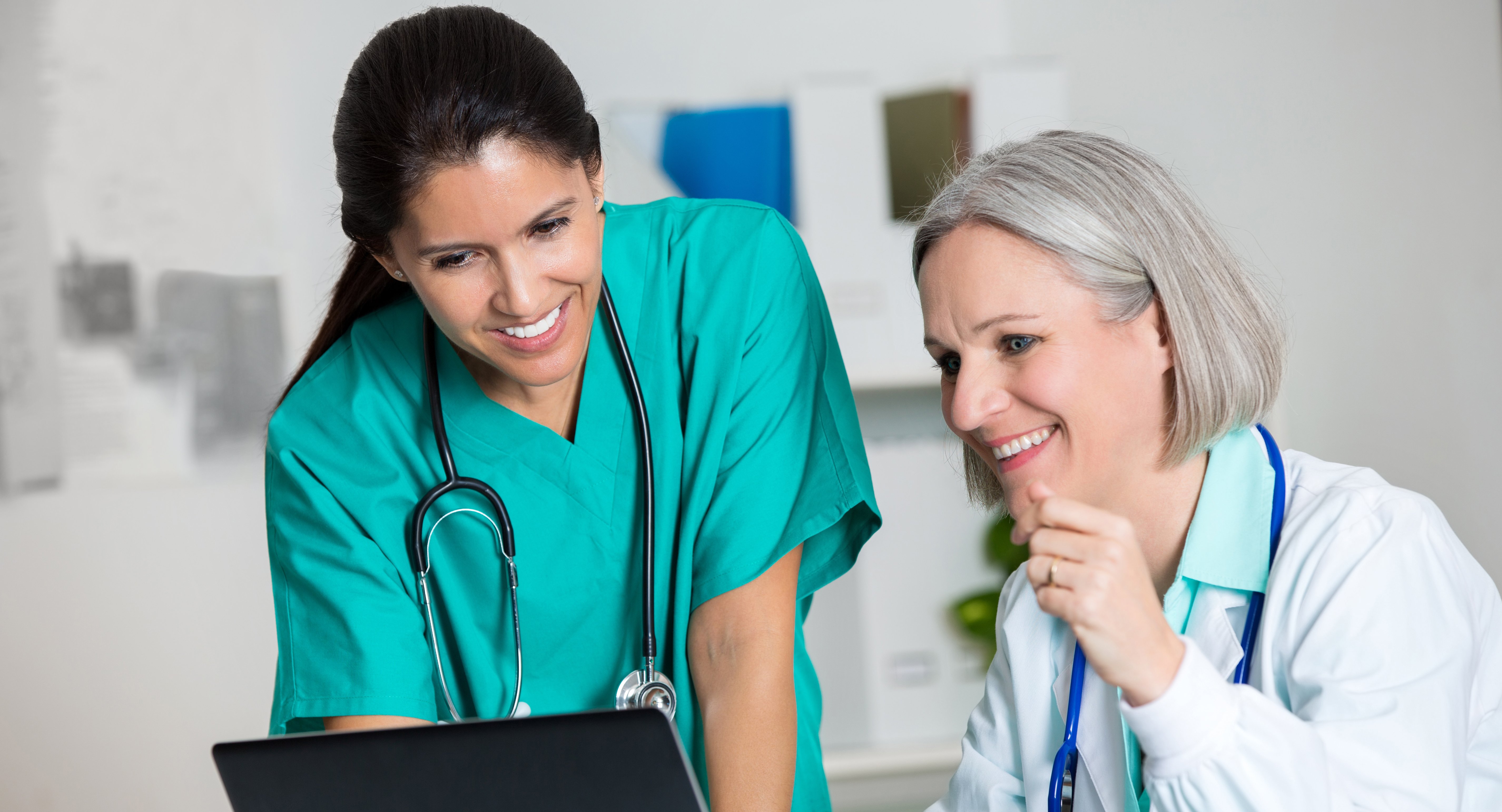 Das Bild zeigt eine sitzende etwas ältere Frau in weißem Kittel mit Stetoskop vor einem Rechner und eine zweite Frau in grünem Kasak mit stetoskop stehend nach vorn gebeugt daneben. Sie schauen zusammen auf den Bildschirm und lächeln.