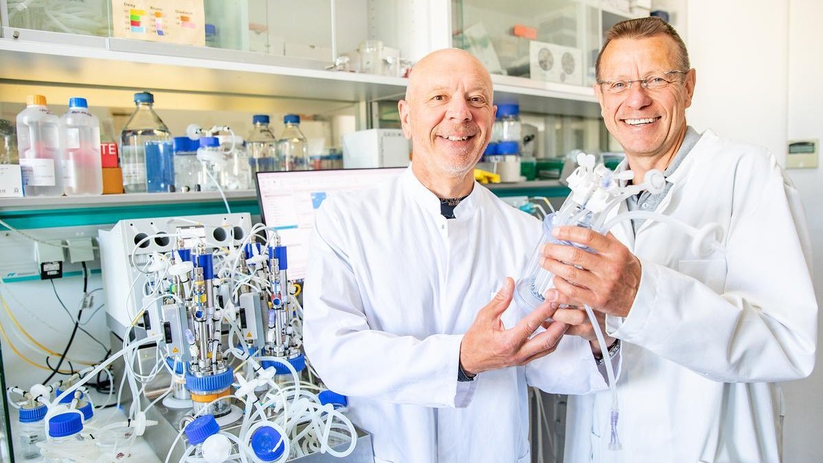 Zwei Forscher in weißen Kitteln stehen in einem Labor und halten einen Bioreaktor in den Händen. 
