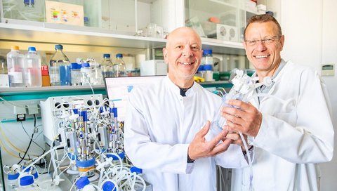 Two scientists in white lab coats are standing in a laboratory, holding a bioreactor in their hands.