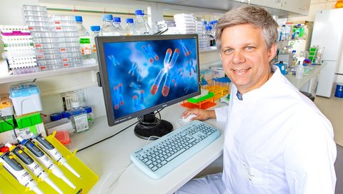The photo shows Prof. Dölken in a white lab coat, sitting on a chair in a laboratory with a screen on the table next to him.