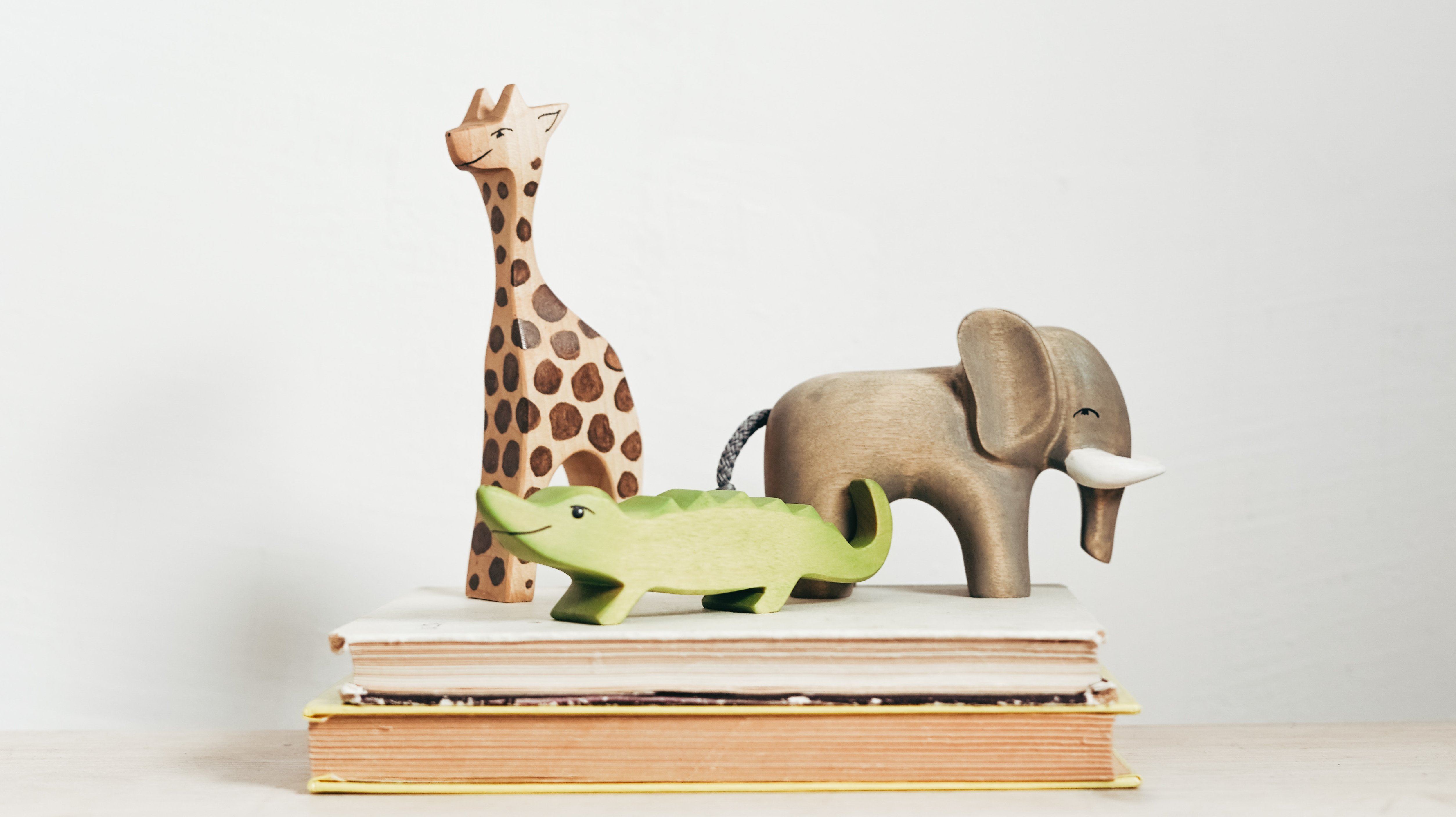 Das Bild zeigt drei freundliche blickende Holzspielfiguren. Eine Giraffe, einen Elefanten und einen grünen Drachen, die auf zwei zugeklappten Büchern stehen.