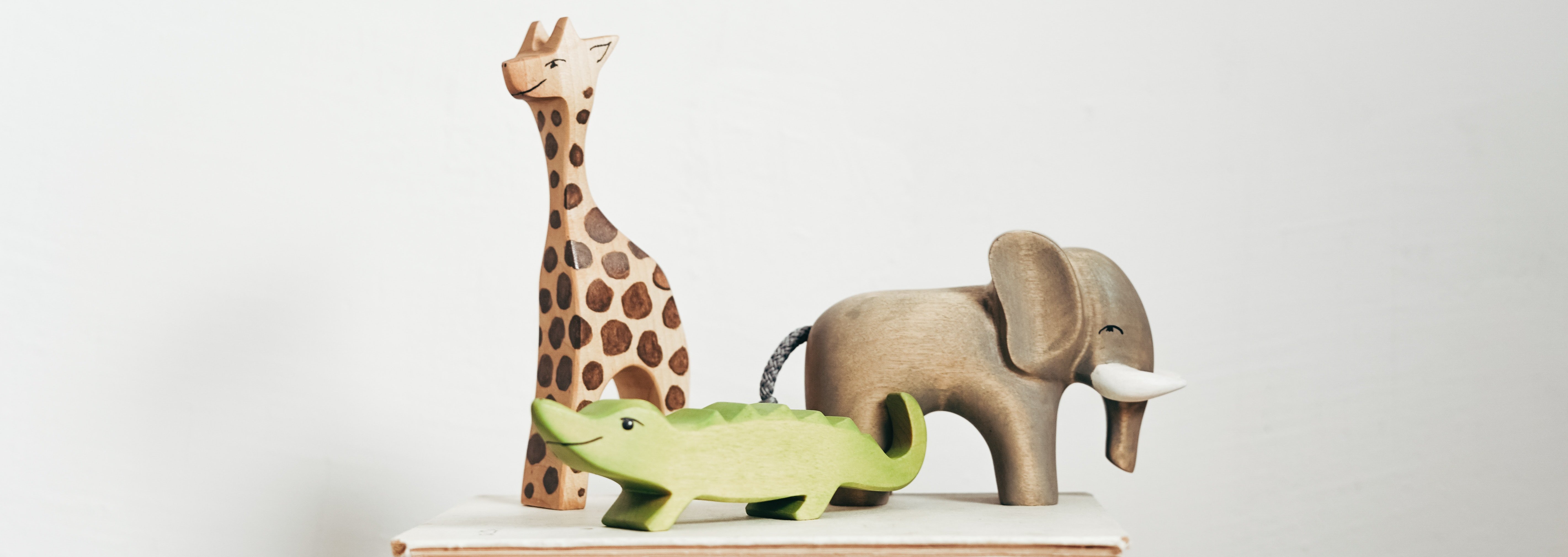 Das Bild zeigt drei freundliche blickende Holzspielfiguren. Eine Giraffe, einen Elefanten und einen grünen Drachen, die auf zwei zugeklappten Büchern stehen.