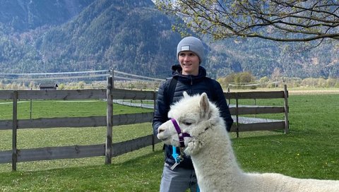 Junger Mann steht mit Alpaka auf einer Wiese. 