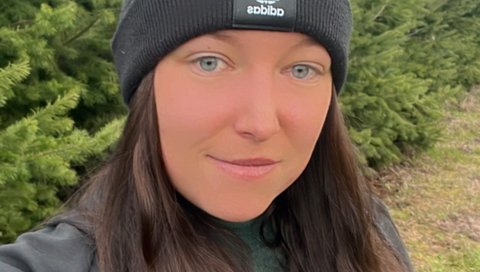 Julia Beese steht vor einem Tannenwald, sie trägt eine schwarzes Oberteil und eine schwarze Mütze, die Haare liegen offen über ihren Schultern. 