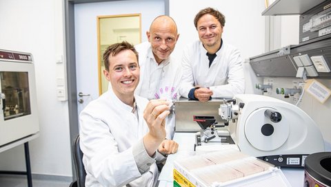 Drei Männer in weißen Kitteln sind nebeneinander zu sehen. Sie schauen in die Kamera und lächeln.