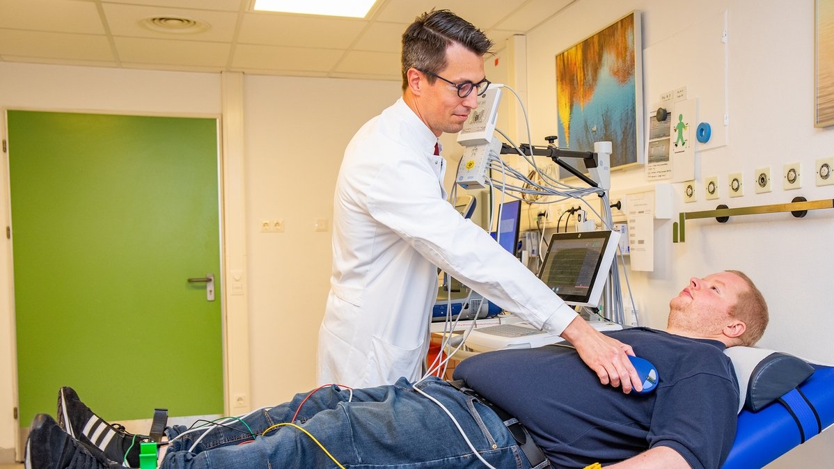 Professor Duncker sits at the bedside of a male patient and checks whether the implanted device is working.