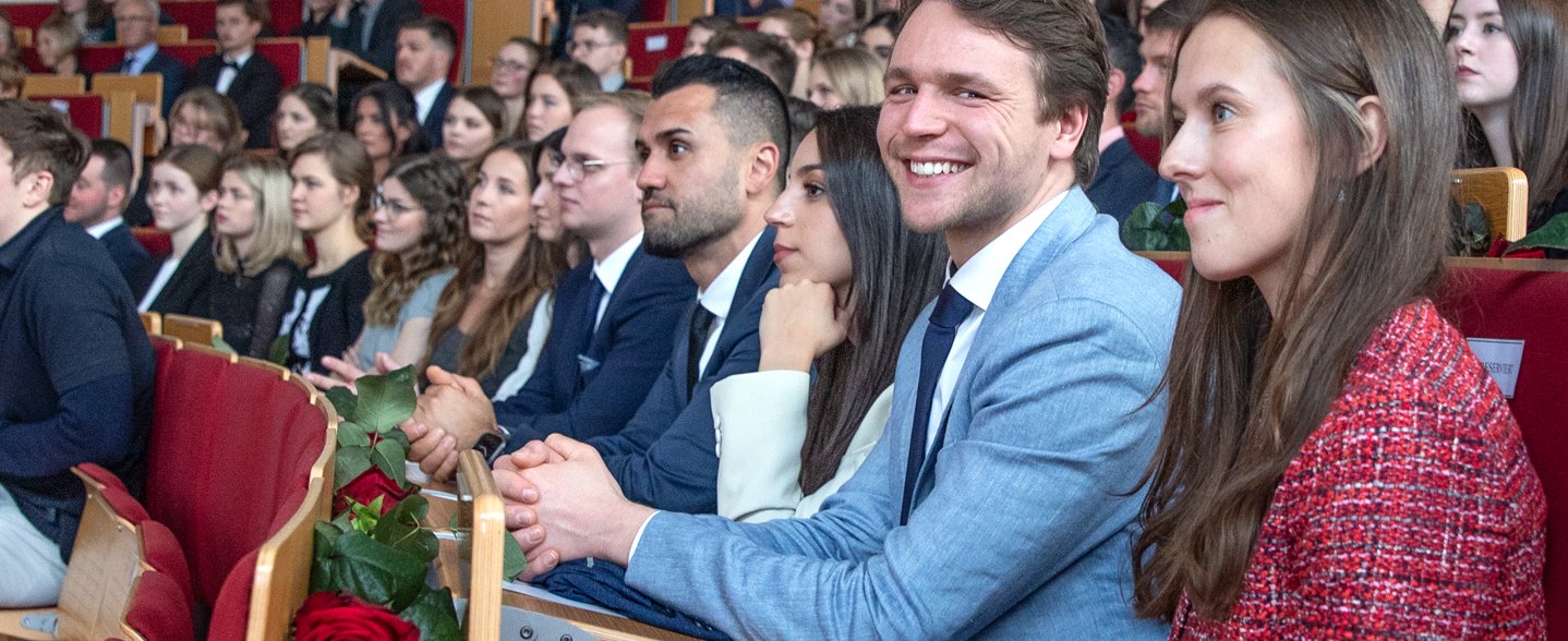 in einem Hörsaal sitzen die Studierenden. Vorne schaut ein Mann in die Kamera und lächelt.