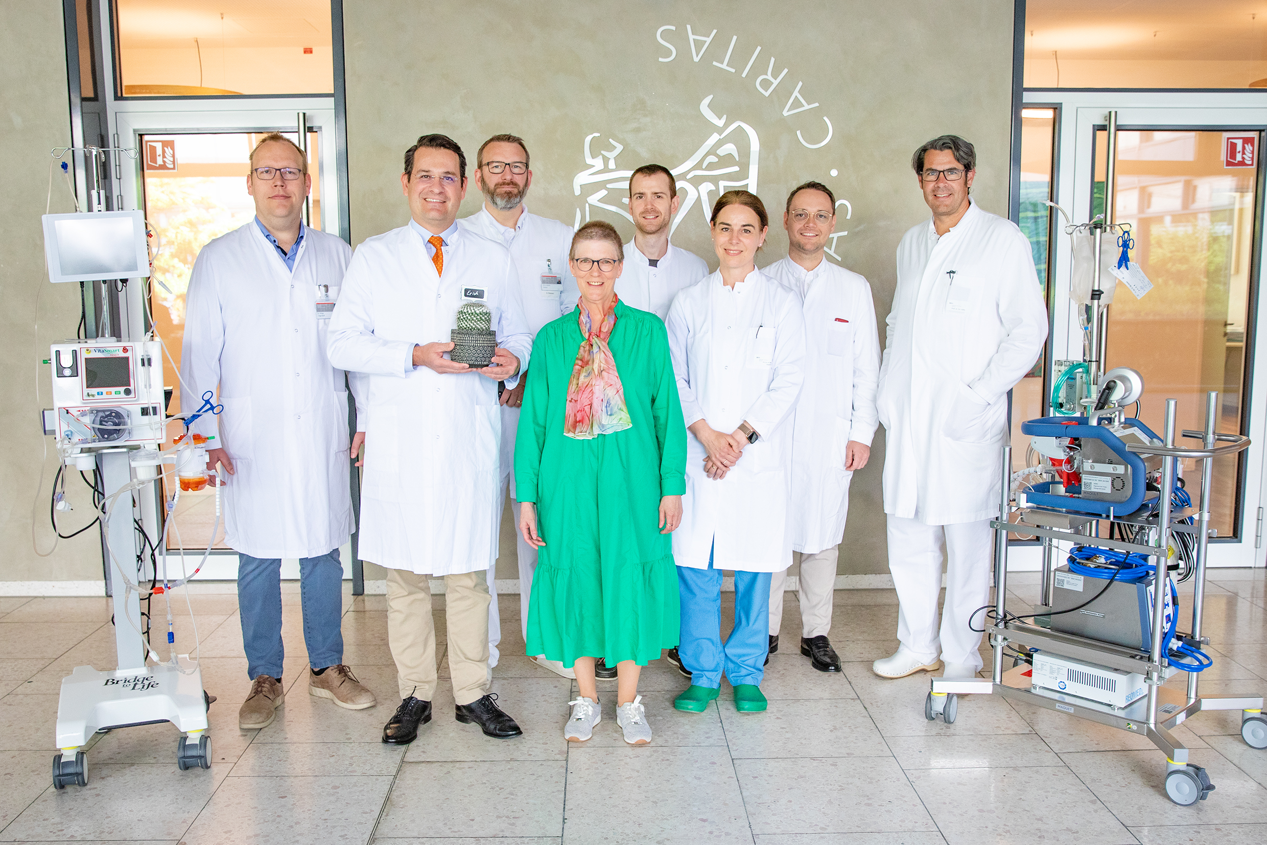 6 Ärzte und eine Ärztin im weißen Kittel sowie die Patientin in der Mitte mit den Geräten, die weährend der OP eingesetzt wurden vor dem Logo der MHH