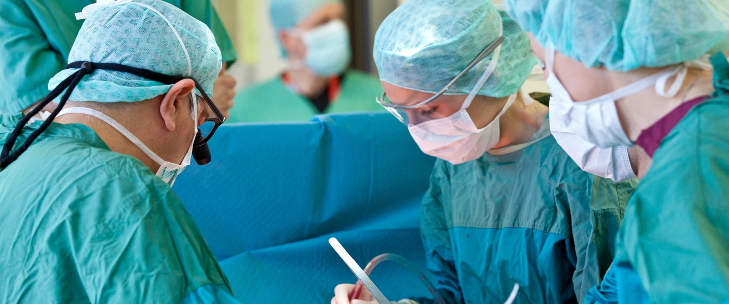 Drei Ärzte stehen in Schutzkleidung am OP-Tisch mit Instrumenten in der Hand. Im hinteren Bereich steht der Anästhesist, abgetrennt von den Chirurgen mit einem grünen Tuch.