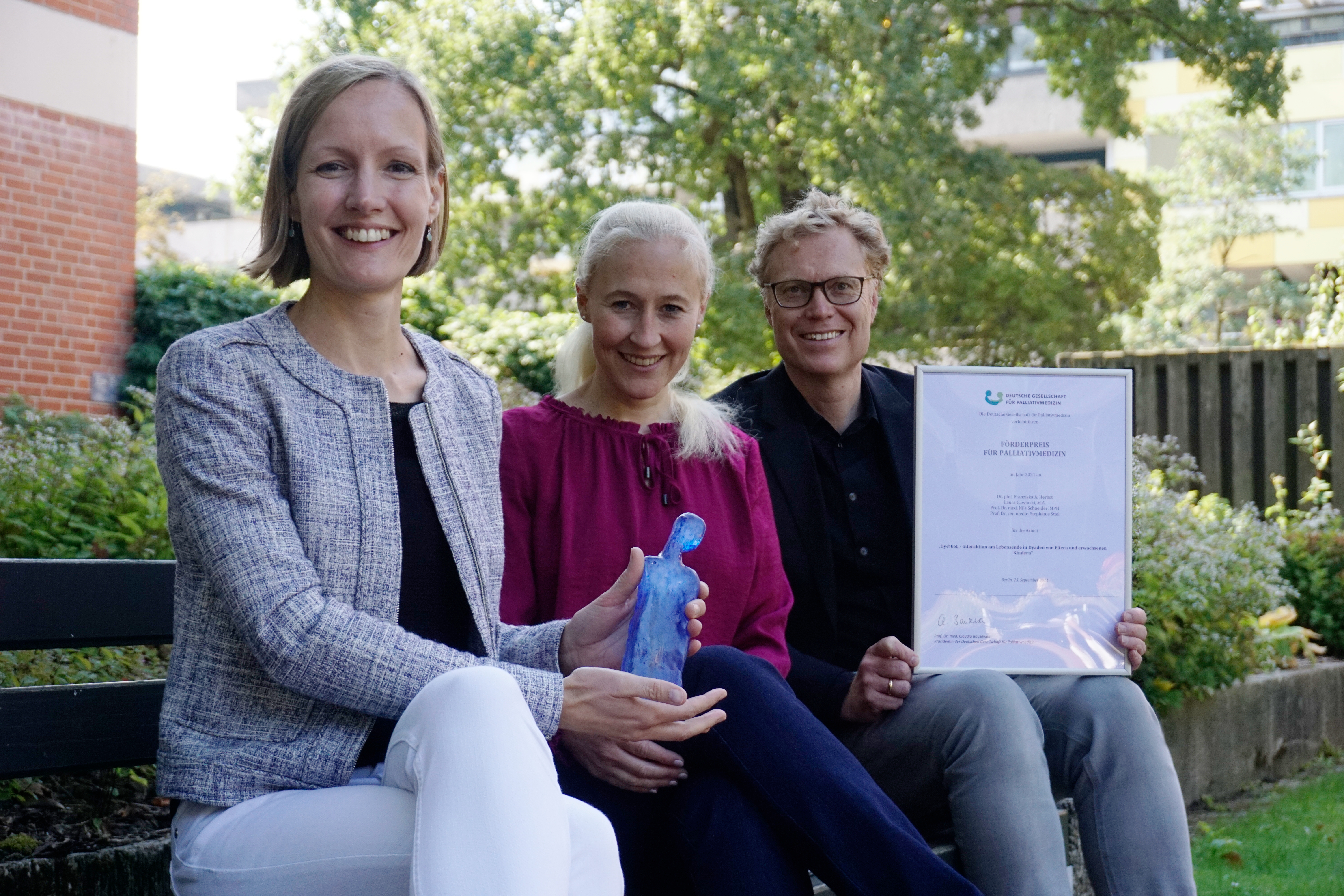 Dr. phil. Franziska Herbst, Professorin Dr. Stephanie Stiel und Professor Dr. Nils Schneider sitzen draußen zusammen auf einer Bank und halten Preisurkunde und -statue in den Händen.