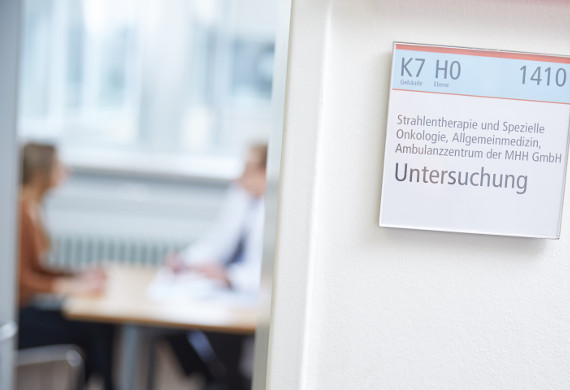 Auf der rechten Bildhälfte sind ein Türrahmen und eine Wand zu sehen. An der Wand hängt ein Schild auf dem „Untersuchung“ steht. Auf der linken Bildseite ist verschwommen im Hintergrund ein Gespräch zwischen einem Mann in einem weißen Kittel—vermutlich einem Arzt—und einer Frau in einem braunen Oberteil—vermutlich eine Patientin—zu sehen.