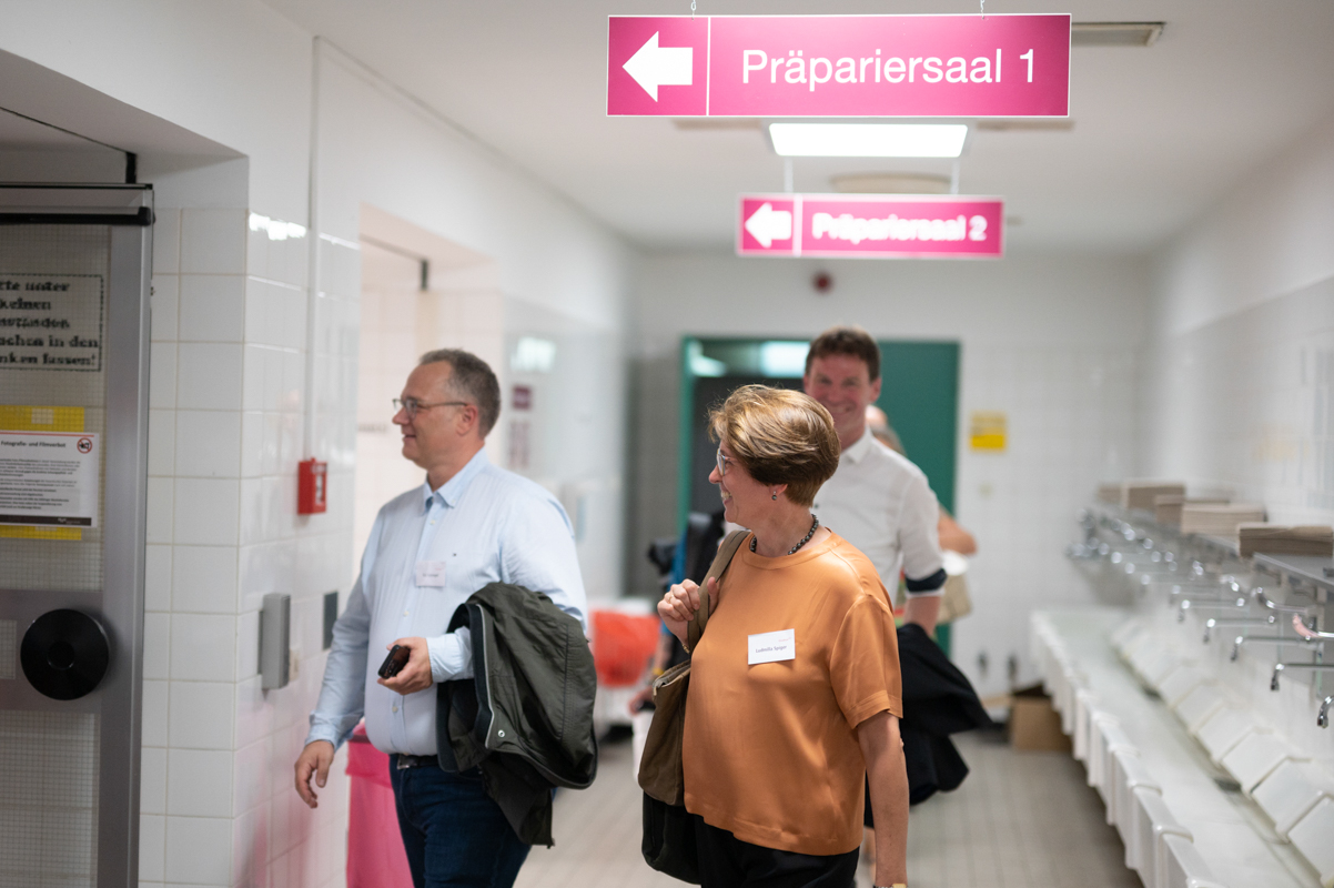 Zwei Absolvent:innen stehen kurz vorm Betreten des Präpariersaals unter einem pinkfarbenen Schild mit der Aufschrift "Präparationssaal".