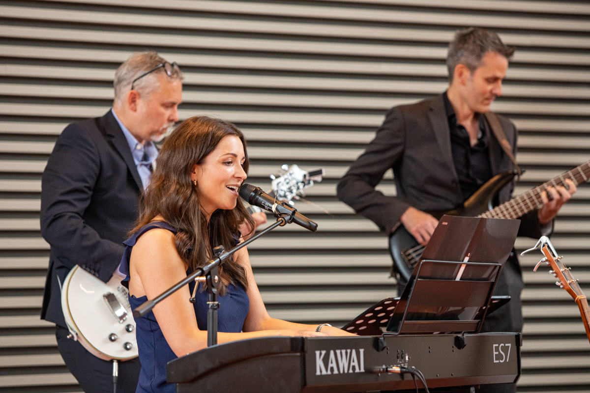 Frau Schulze sitzt am Keyboard und singt. Im Hintergrund sind ein Gitarrist und ein Bassist zu sehen.