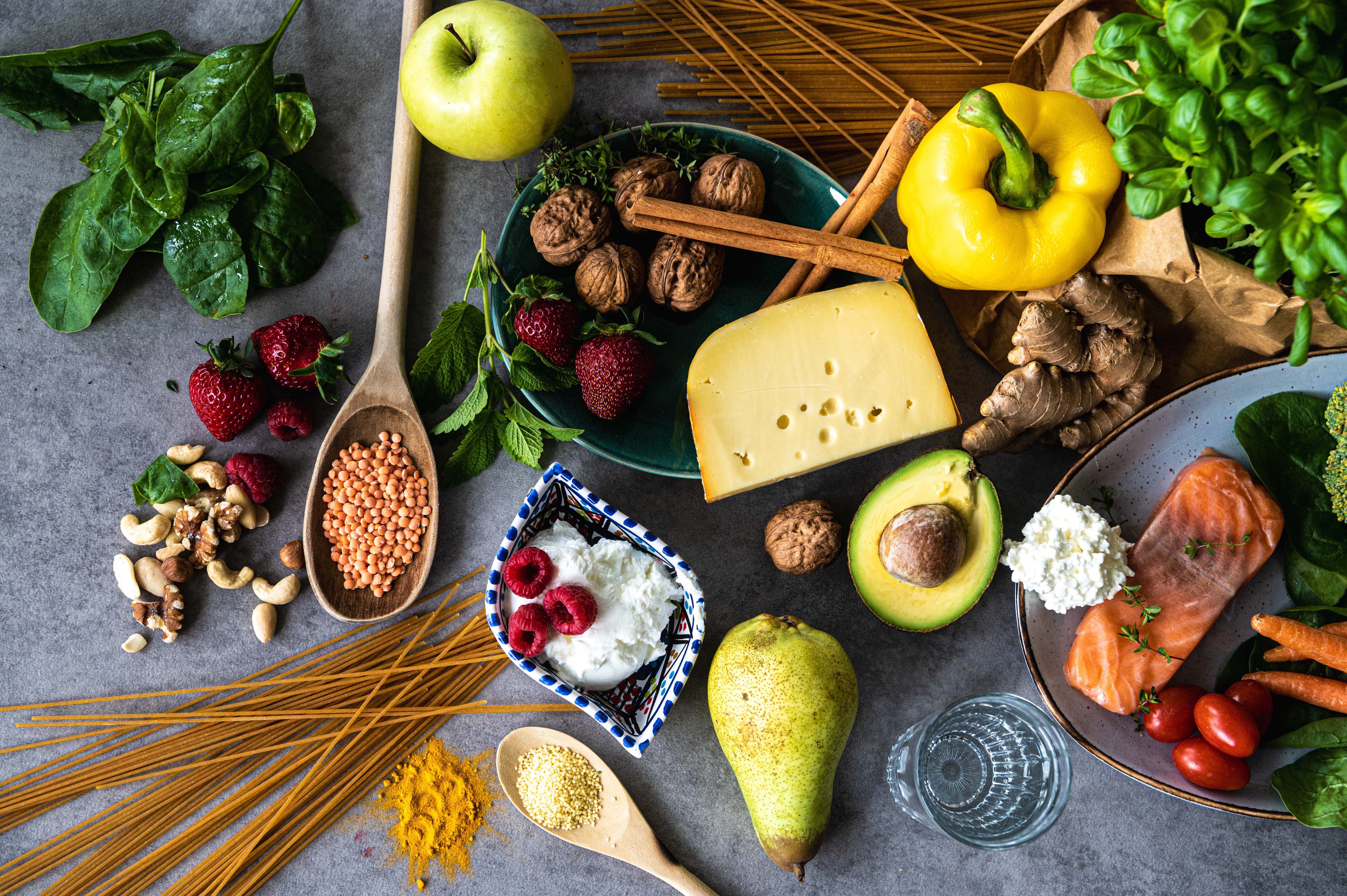 Auf einem grauen Hintergrund liegen verschiedene Lebensmittel darunter Käse, Nudeln, Linsen, Obst, Gemüse, Fisch und Nüsse sowie Kräuter.
