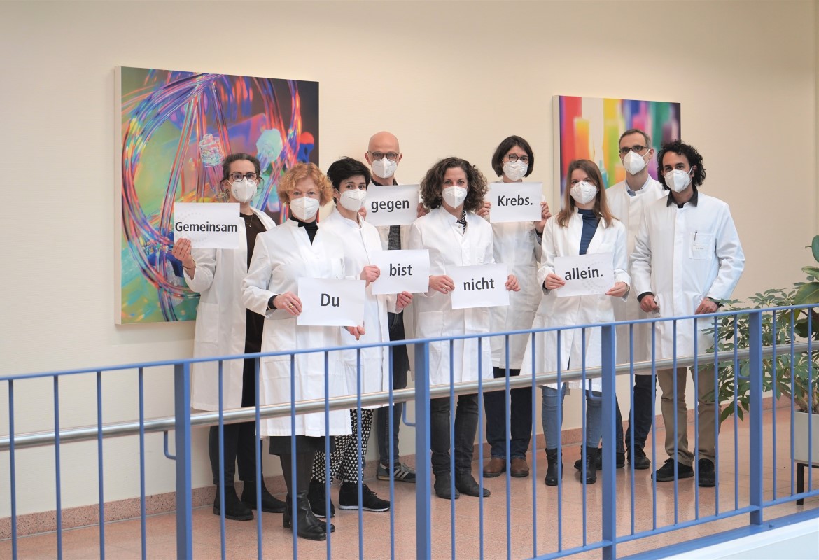 Mitarbeiterinnen und Mitarbeiter aus dem Team Humangenetik in weißen Kitteln halten ein Schild hoch, auf dem steht "Gemeinsam gegen Krebs. Du bist nicht allein."
