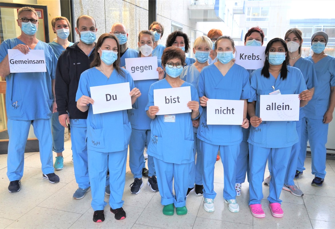 Das Pflegeteam in blauer Kleidung hält Schilder hoch, auf denen steht: "Gemeinsam gegen Krebs. Du bist nicht allein.". 