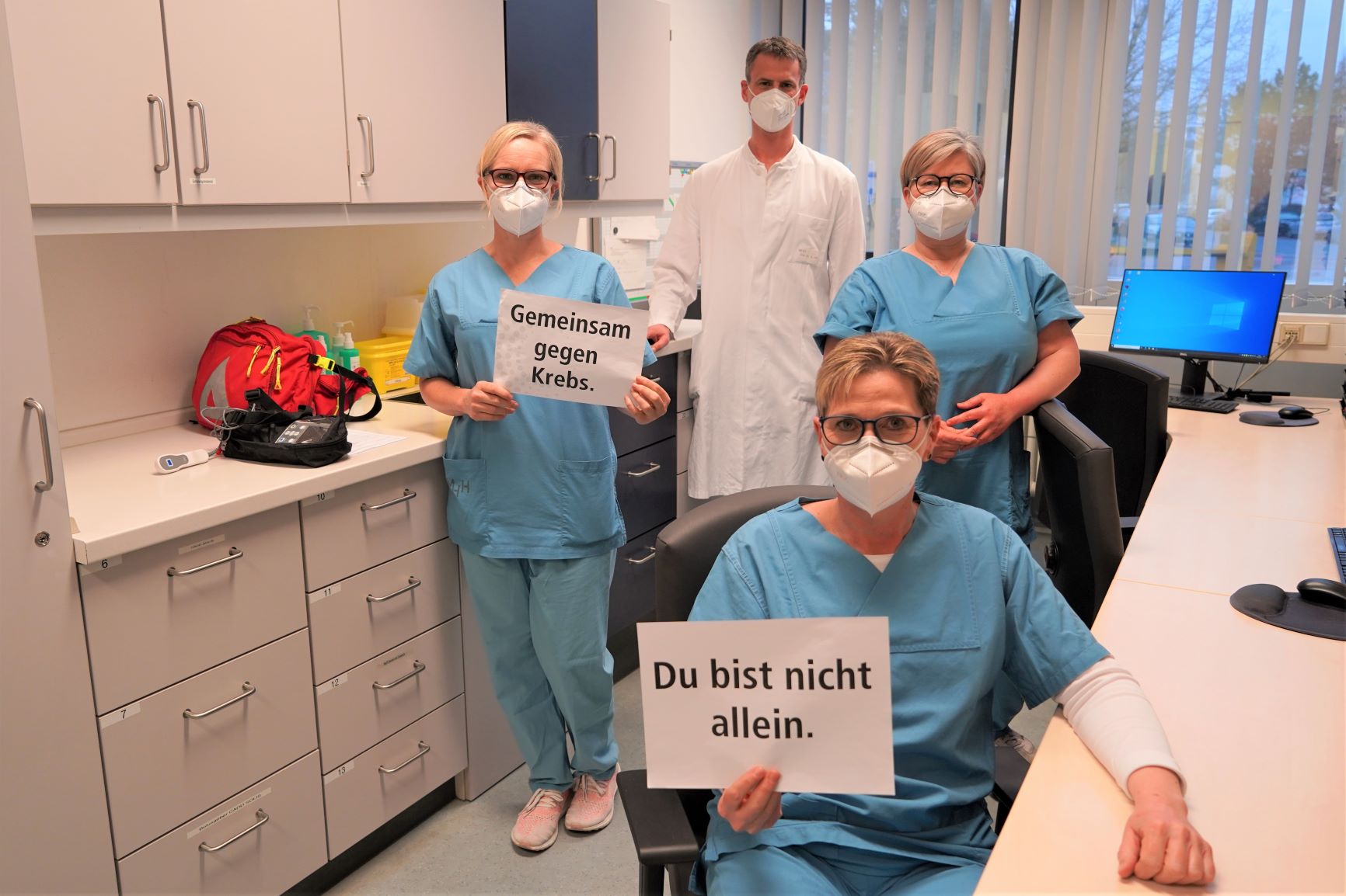 Vier Mitarbeiter vom Schmerzdienst im Stationsraum halten ein Schild, auf dem steht "Gemeinsam gegen Krebs. Du bist nicht allein."