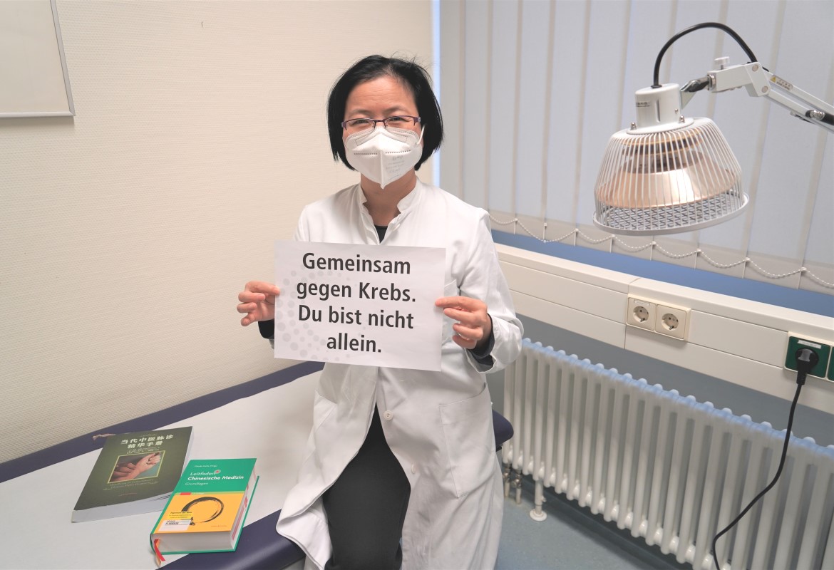 Frau Dr. Zheng aus dem Institut für Traditionelle Chinesische Medizin sitz im weißen Kittel auf einer Behandlungsliege und hält ein Schild hoch, auf dem steht: "Gemeinsam gegen Krebs. Du bist nicht allein.". 