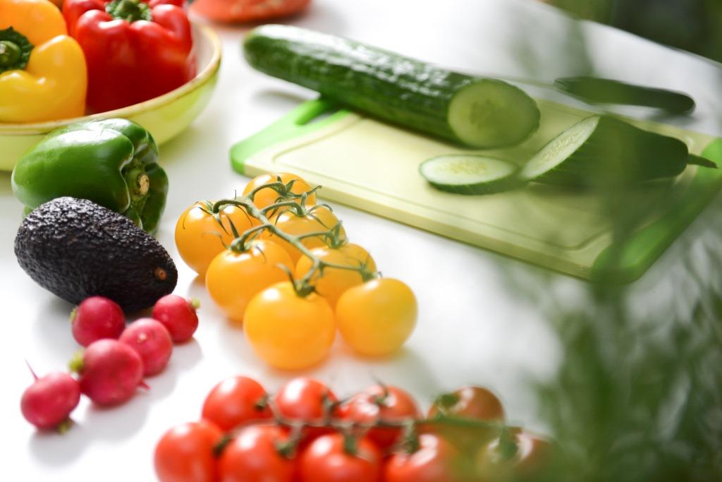 Gemüse wie eine Gurke, Paprika und Tomaten liegen auf einem weißen Tisch. Daneben ein Schneidebrett mit Messer.