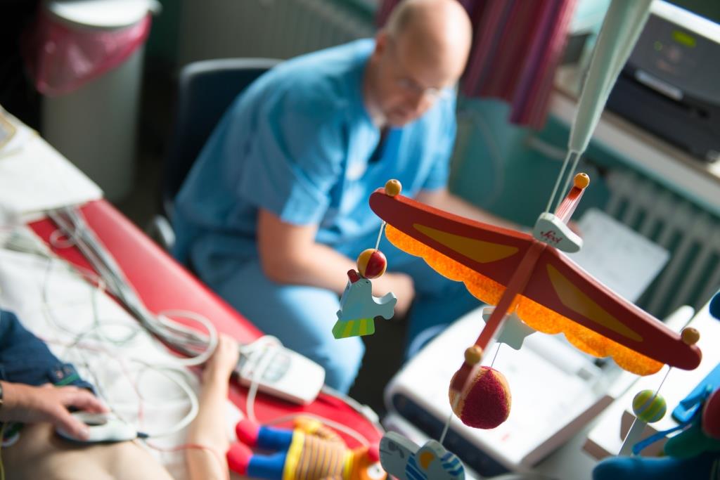 Ein Krankenpfleger aus der Vogelperspektive in einem Patientenzimmer der neben einem Kinderbett sitzt.