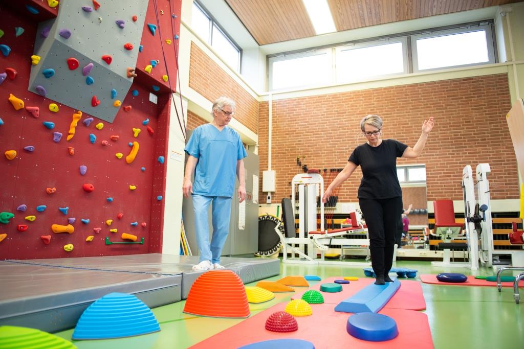 Copyright: medJUNGE Der Rehabilitationsraum der MHH mit verschienden Geräten. Eine Patientin balanciert auf einer trapezförmigen Erhöhung. Neben ihr läuft eine anleitende Person.