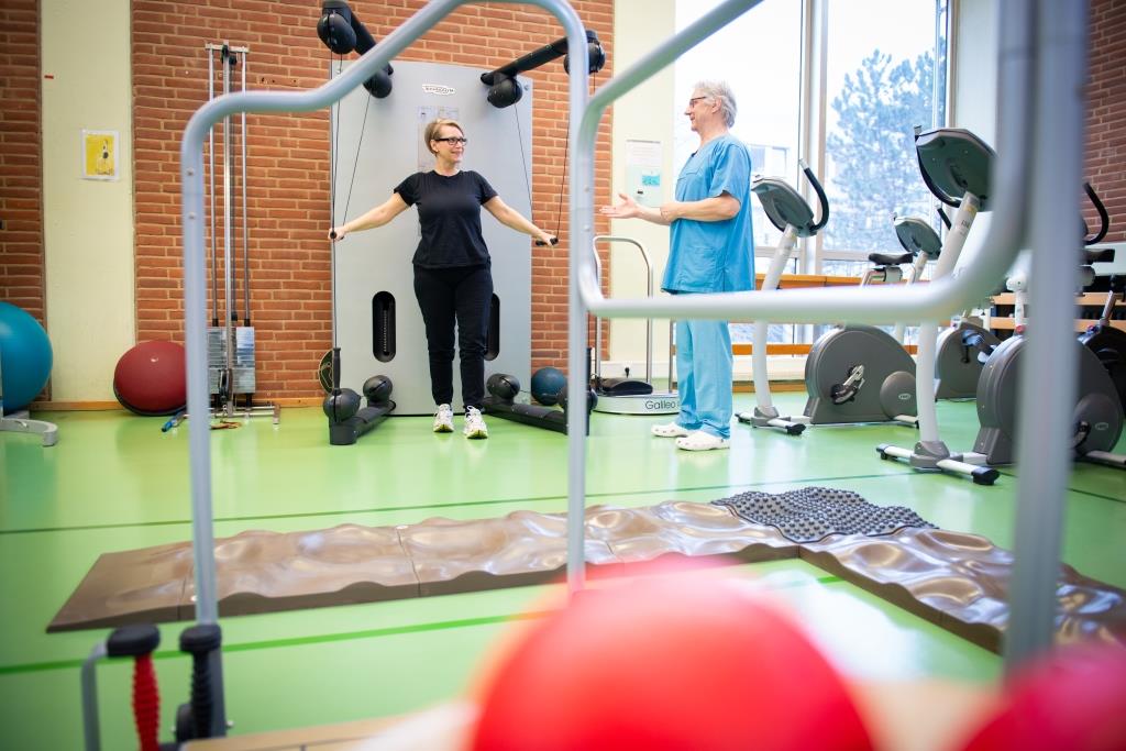 Copyright: medJUNGE Der Rehabilitationsraum der MHH mit verschienden Geräten. Eine Patientin führt eine Oberkörperübung an einem Gerät aus. Neben ihr steht eine anleitende Person.