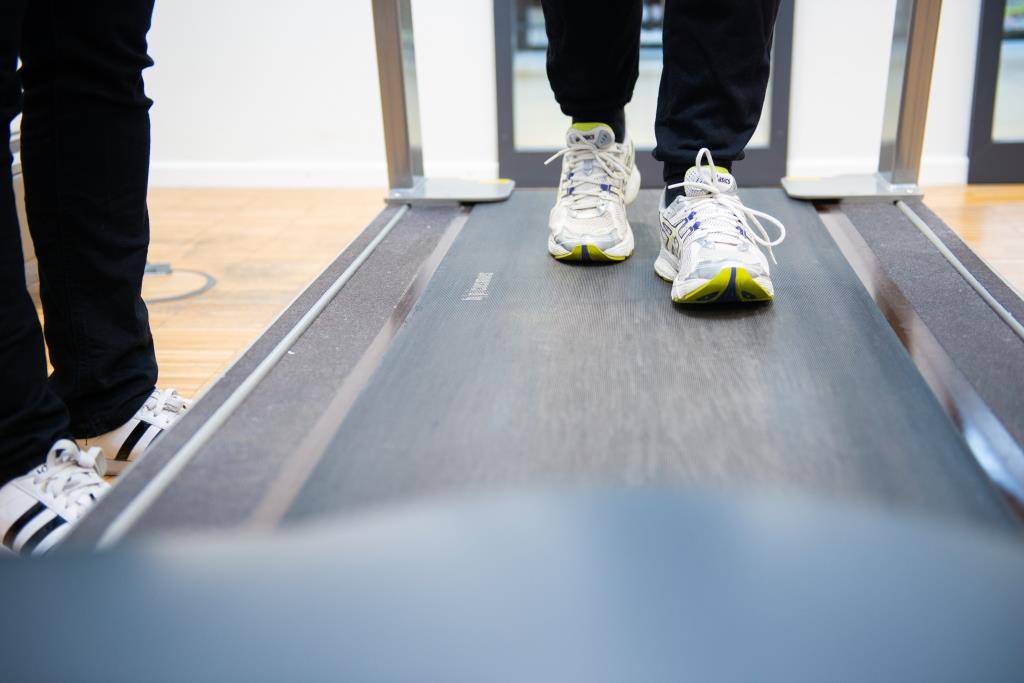 Eine Person geht mit Turnschuhen auf einem Laufband. Neben ihr steht eine anleitende Person.