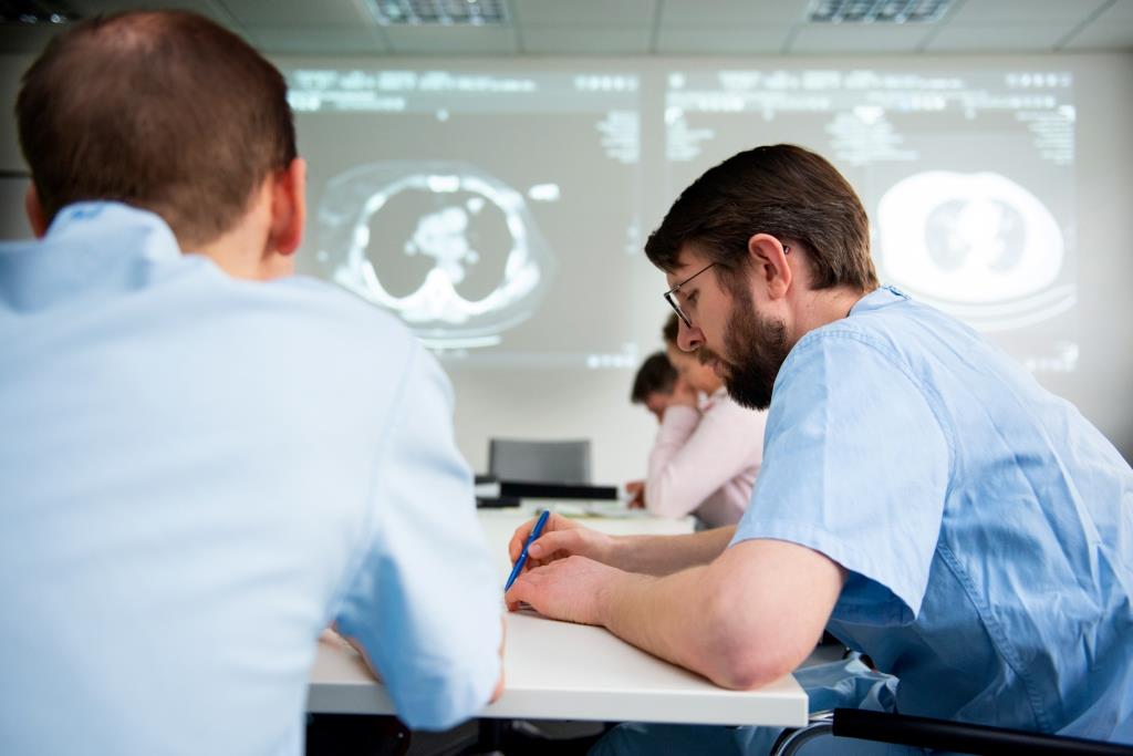 Nahaufnahme von zwei Ärzten die am Tisch in einer Tumorkonferenz sitzen. Im Hintergrund sind CT Bilder zu sehen.