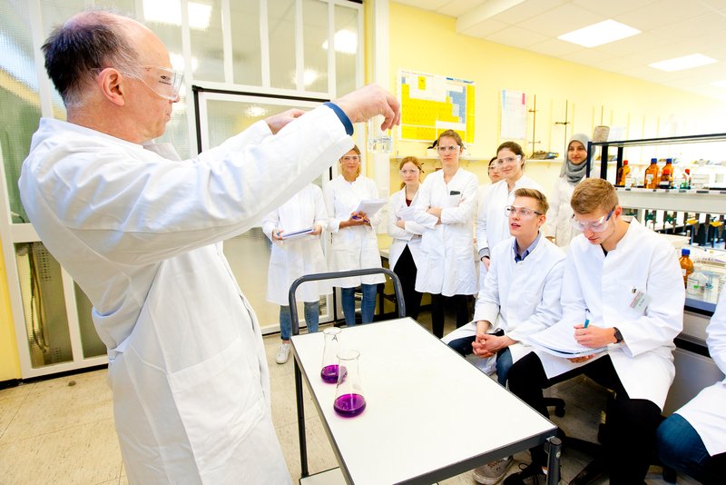 Ein Dozent im weißen Kittel der seinen Studierenden im Chemieraum etwas veranschaulicht.