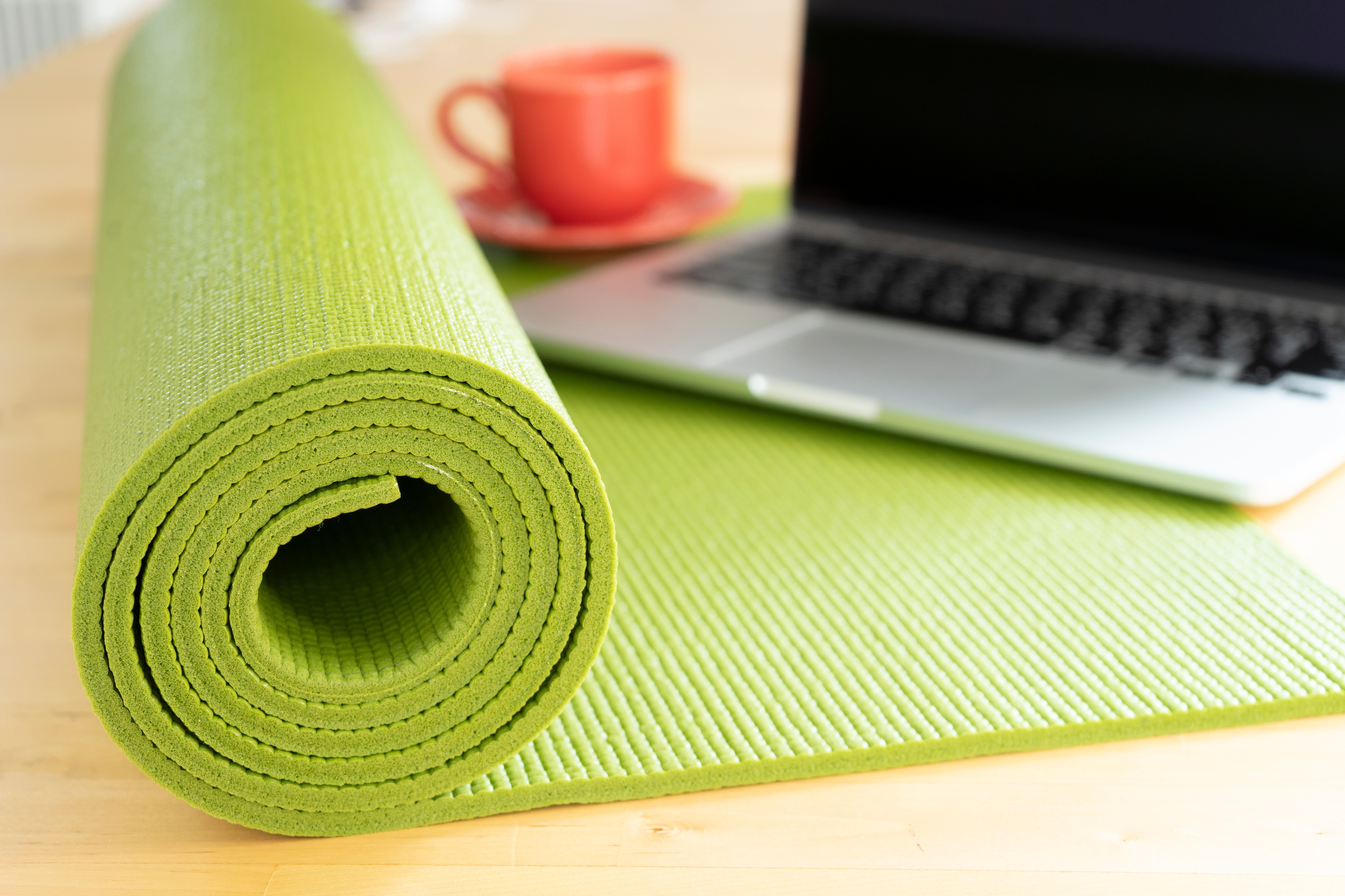Eine grüne Yogamatte liegt auf dem Schreibtisch. Daneben steht ein Laptop sowie eine rote Tasse.