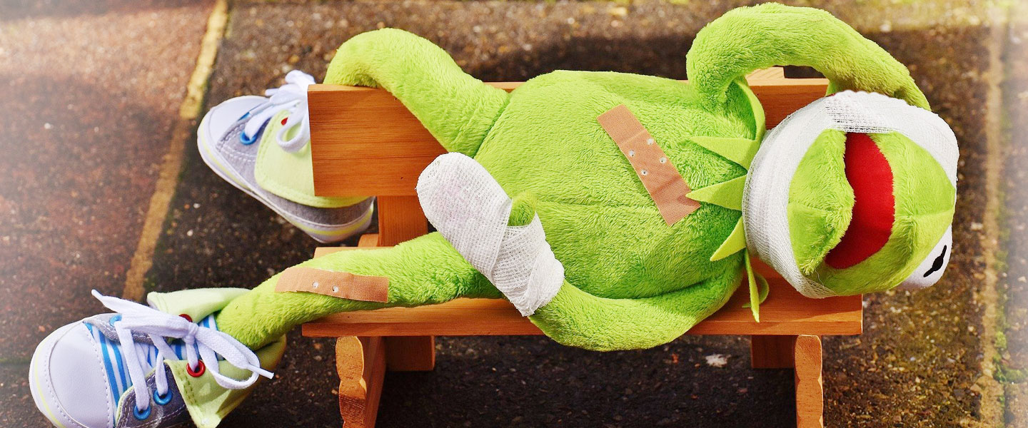 grüner Stofffrosch liegt auf einer Bank und hält sich den verbundenen Kopf