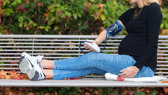 eine Schwangere sitz auf einer Bank mit Blutdruckmessgerät in der Hand