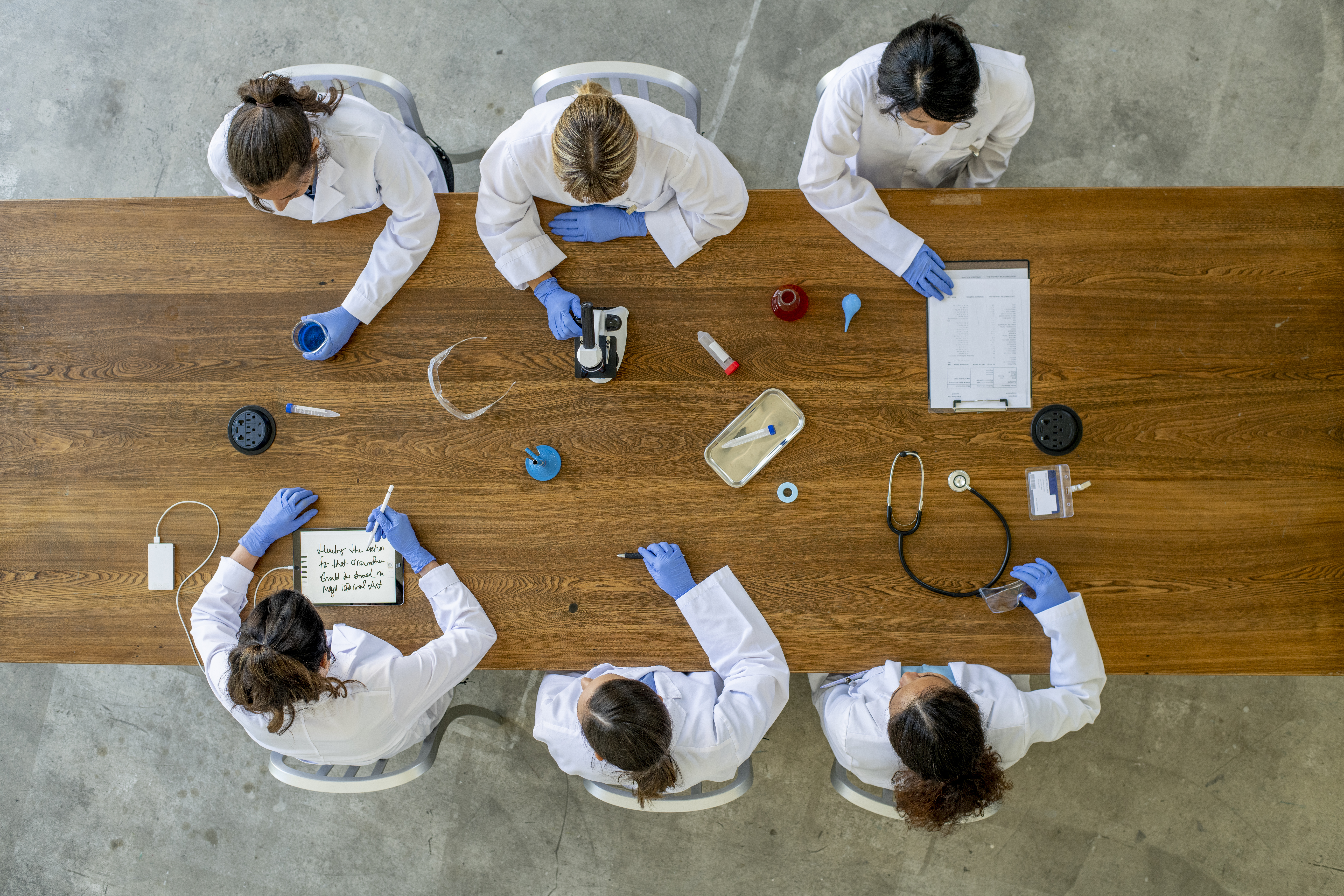 An einem Holztisch stitzen sechs Frauen in weißem Kittel, vor ihnen liegen verschiedene Dinge auf dem Tisch, wie Stetoskop, Pipeten, Mikroskop). Das Foto wurde von oben aufgenommen.