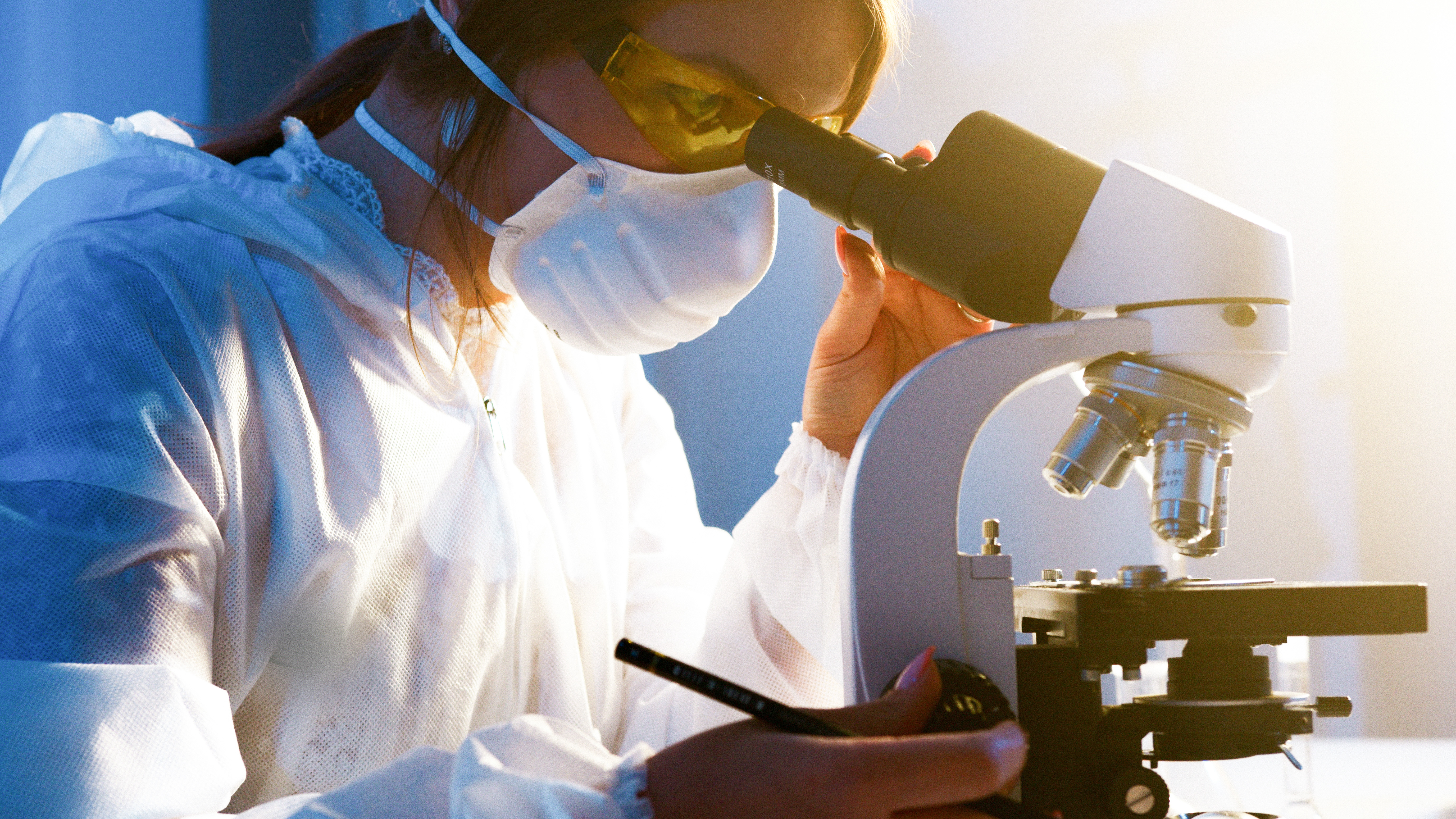 Das Bild zeigt eine Frau in Schutzkleidung, die durch ein Mikroskop schaut und in der rechten Hand einen Stift hält.
