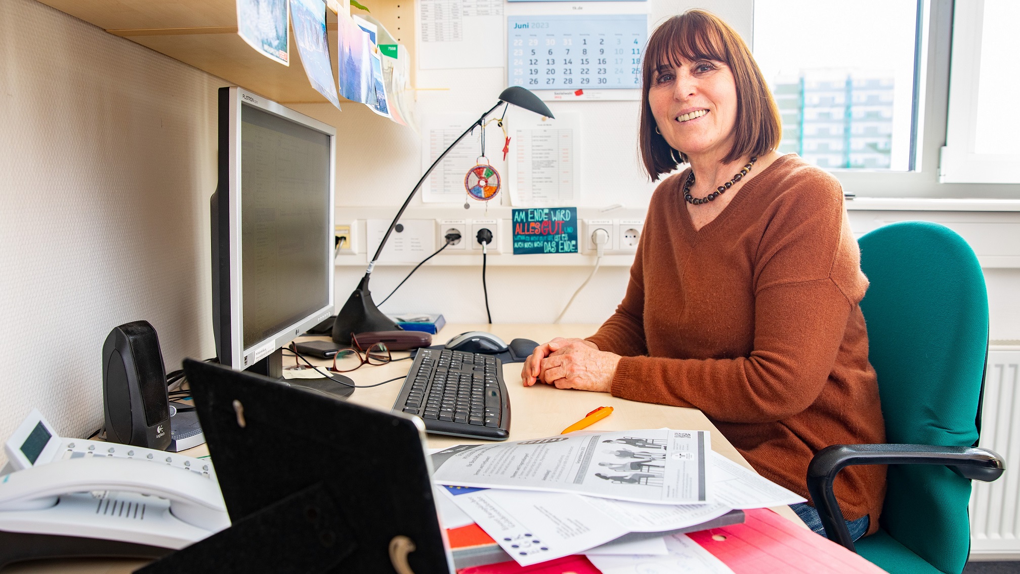 Iris Winter sitzt an ihrem Schreibtisch in der MHH. Darauf zu sehen sind ein PC-Monitor, eine Tastatur, ein Bilderrahmen und mehrere Blätter Papier. 