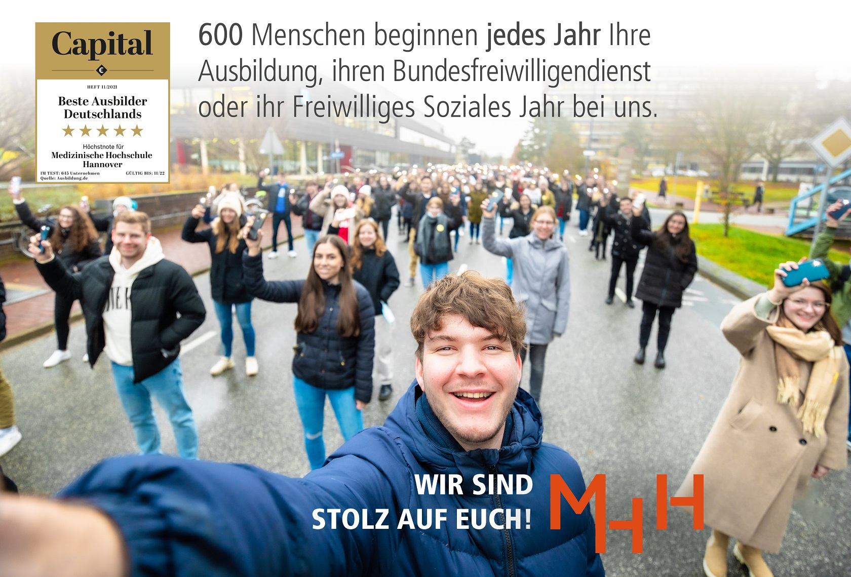 Viele junge Menschen stehen in mehreren Reihen hintereinander auf einer Straße und halten ihre Handys hoch. Oben links ist ein Logo für den besten Ausbilder eingeblendet. Schrift in schwarz: 600 Menschen beginnen jedes Jahr ihre Ausbildung, ihren Bundesfreiwillingendienst oder ihr Freiwilliges Soziales Jahr bei uns. Schrift in weiß: Wir sind stolz auf Euch. Logo MHH in rot.