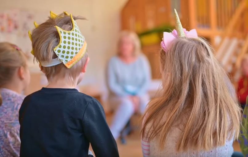 Mehrere Kinder sitzen in einem Stuhlkreis, einige tragen Kopfschmuck wie eine Krone aus Papier und einen Haarreifen mit Einhorn.