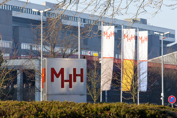 Die Metallstele an der Haupteinfahrt mit den roten Buchstaben MHH. Dahinter ein Gebäude mit drei Fahnen mit MHH-Aufschriften. 