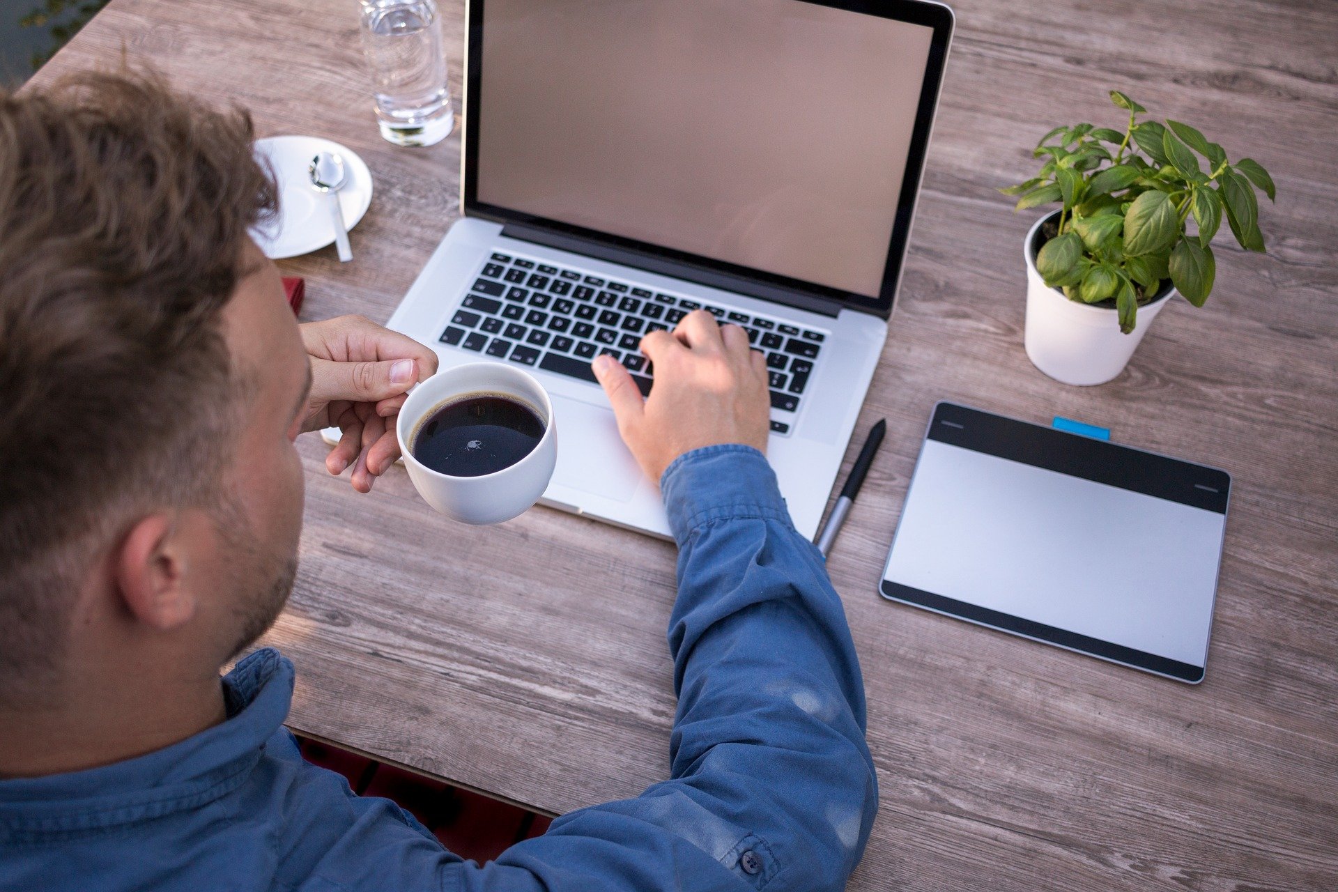 Ein Mann sitzt mit einer Tasse Kaffee in der Hand am Schreibtisch vor einem Laptop
