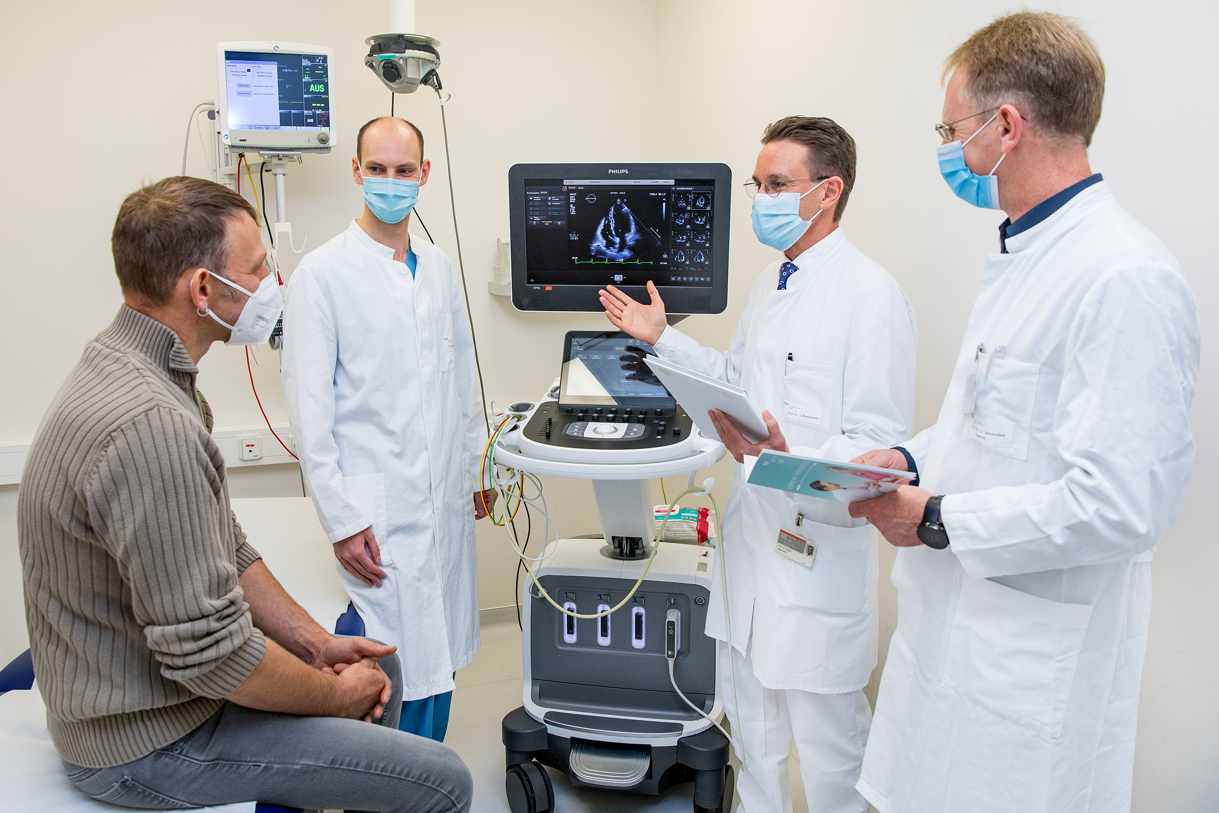Patient Thorsten S. sitzt auf einer Patientenliege, rechts daneben stehen Dr. Fabian Rathje, Professor Dr. Johann Bauersachs und Professor Dr. Udo Bavendiek. In der Mitte ein Bildschirm mit einer Aufnahme des Herzens.  