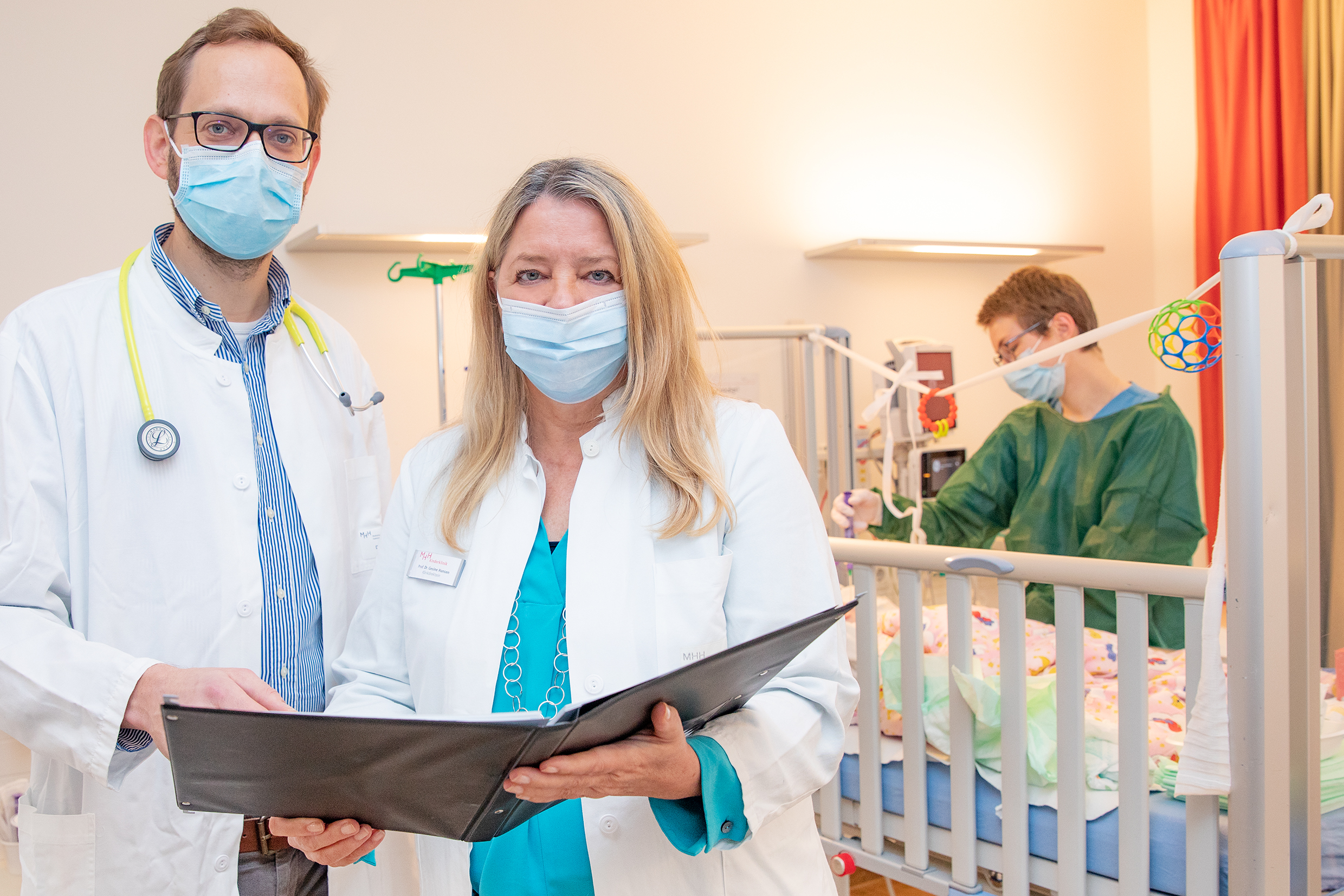 Vor einem Kinderbett in einem Krankenzimmer stehen PD Dr. Martin Wetzke und Professorin Dr. Gesine Hansen. Am Bett kümmert sich eine Pflegerin um ein krankes Kind.