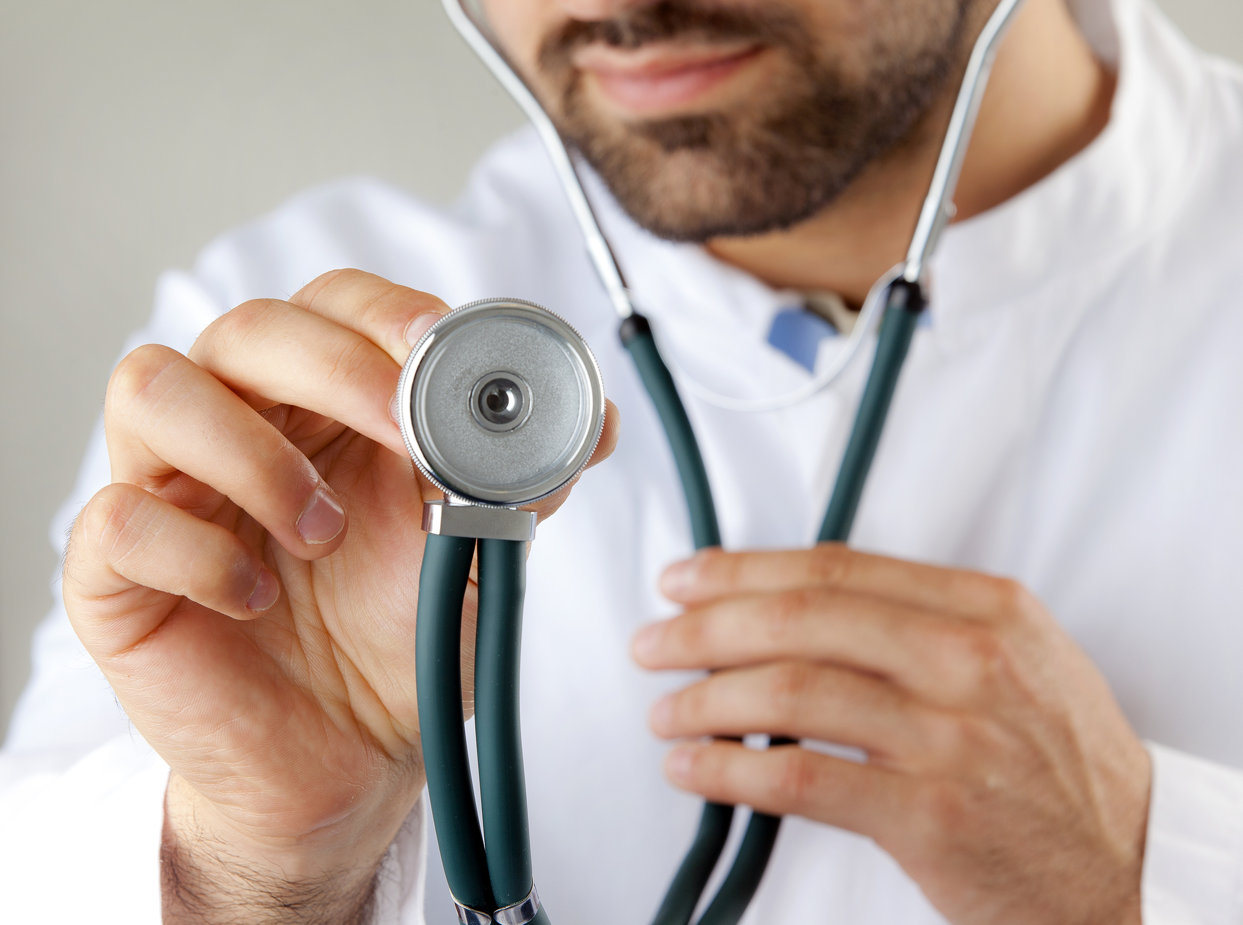 A doctor approaches the viewer with the head of a stethoscope.