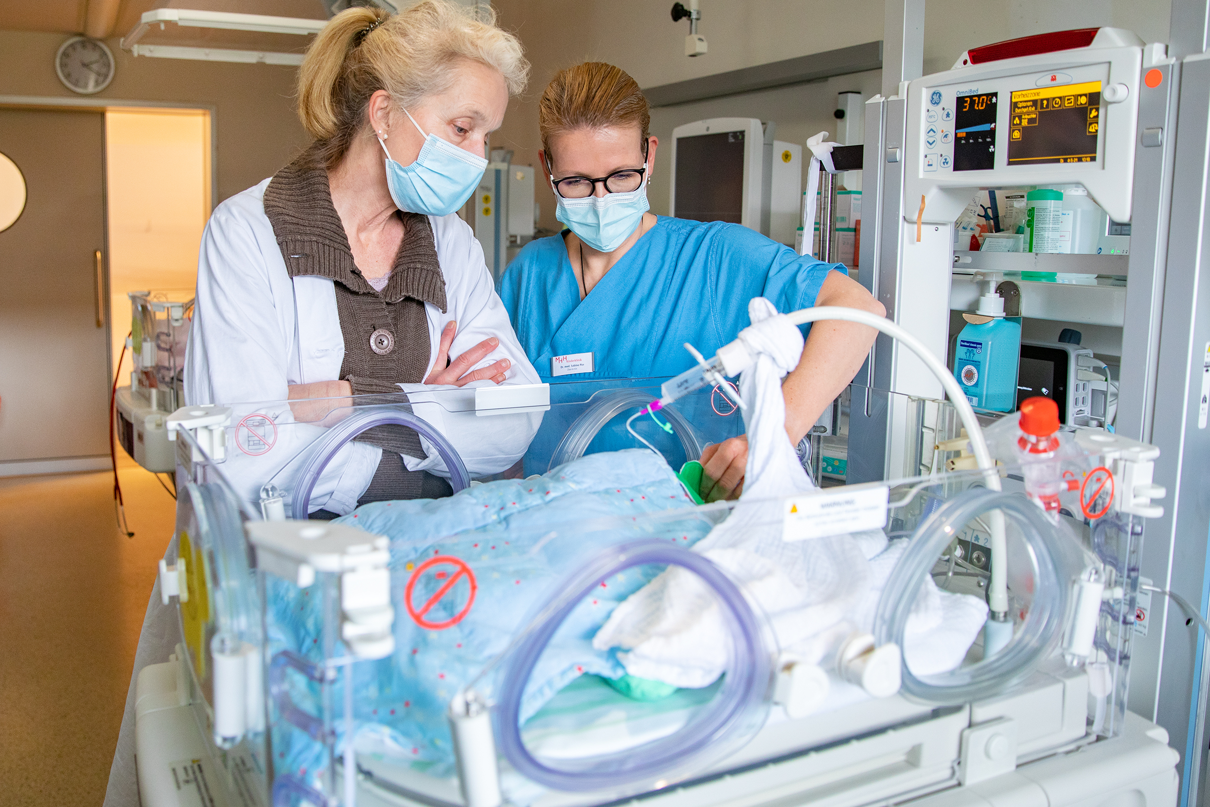 Professor Viemann and PD Dr Sabine Pirr. They are standing at the bedside of a premature baby on the ward for newborns and premature babies at Hannover Medical School, looking in