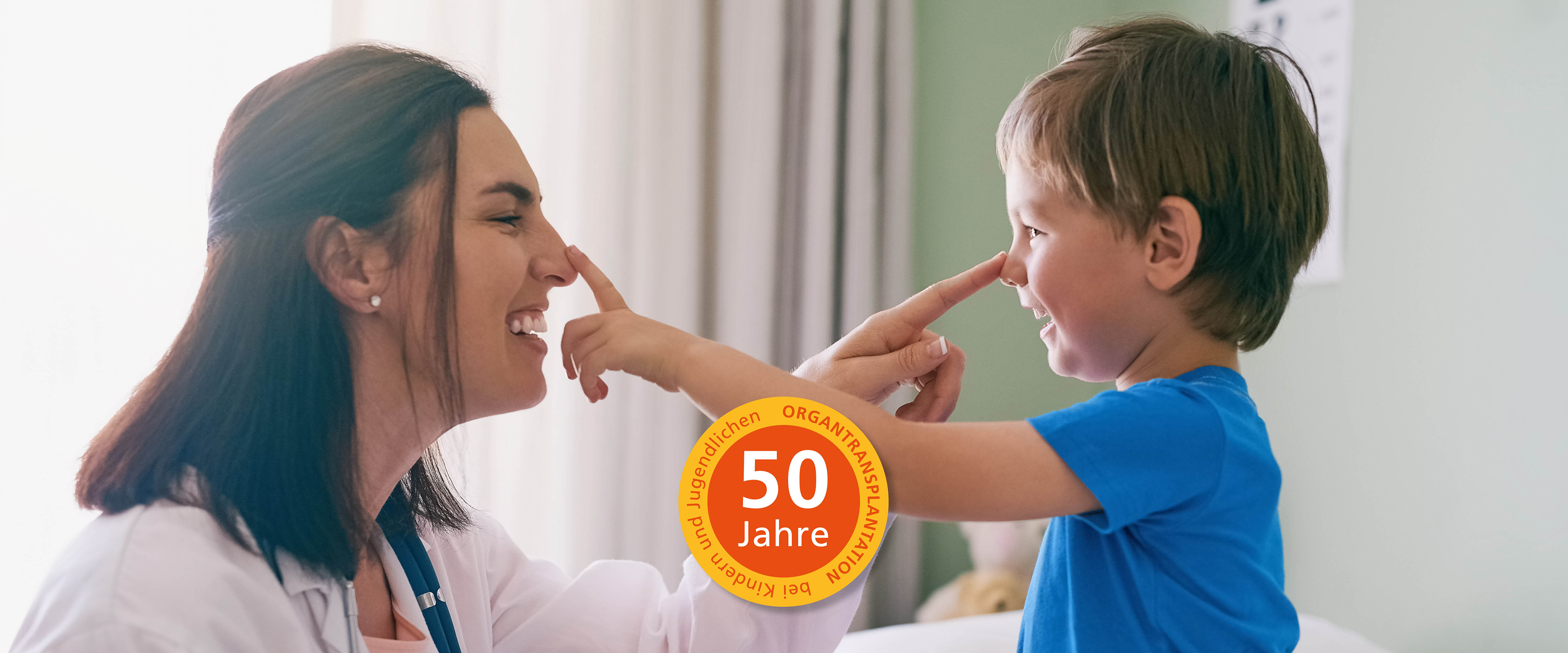 Eine lachende Kinderärztin links im Bild und ein kleiner Junge in blauen T-Shirt tippen sich gegenseitig mit dem Zeigefinger auf die Nase. In der Mitte des Bildes befindet sich ein orange-rotes  Logo zu 50 Jahre Organtransplantation bei Kindern und Jugendlichen